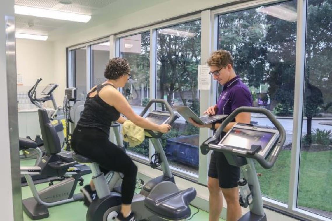 Woman on exercise bike while man stands nearby with laptop. Supplied.