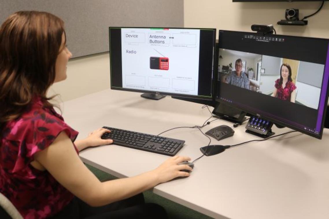 A woman sits at a desk looking at two monitors while she conducts a Zoom call with her face and a man's shown on one of the screens.