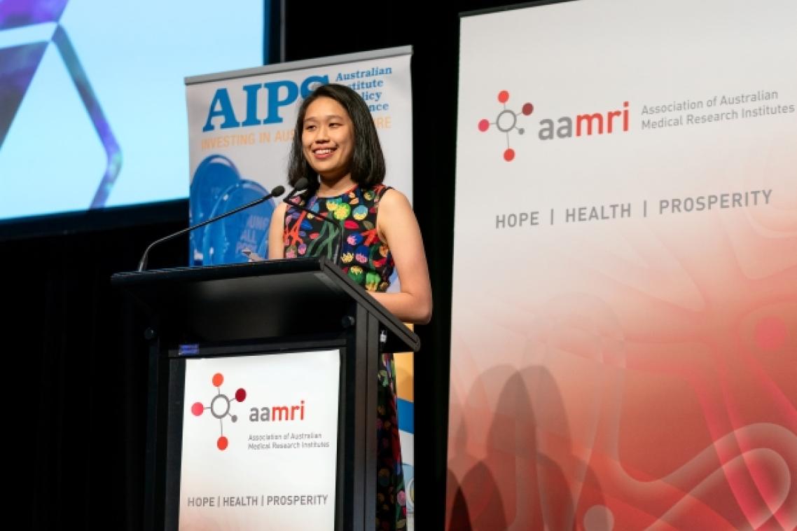 a woman stands at a lectern with banners behind her