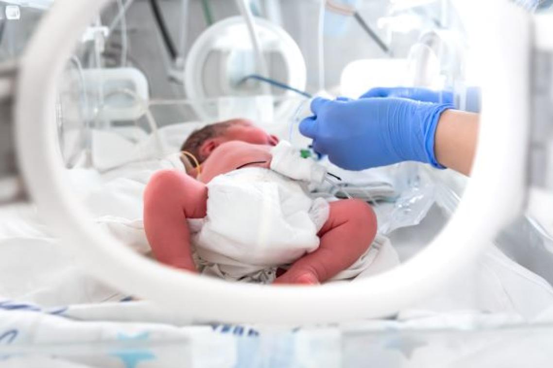 A pre-term baby lies in a humidi-crib while two blue gloved hands administer medicine.