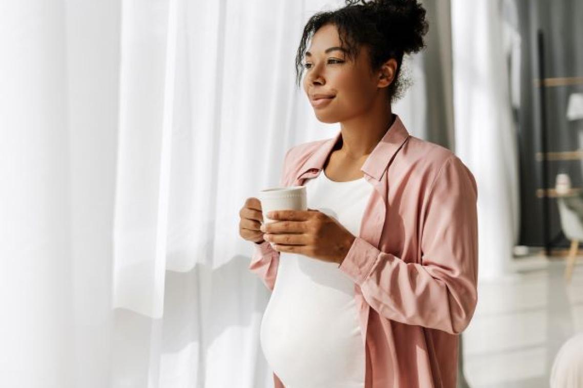 A pregnant woman holds a coffee mug and looks through sheer curtains out a window.