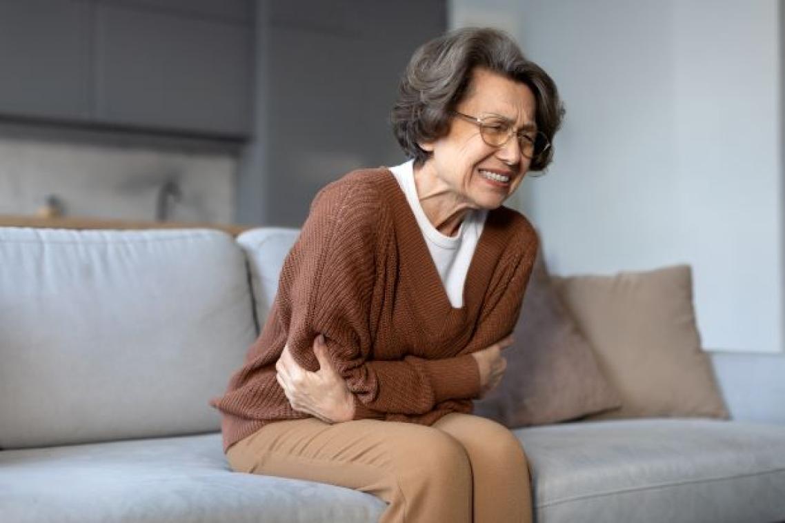 an older woman sits on a sofa clutching her stomach