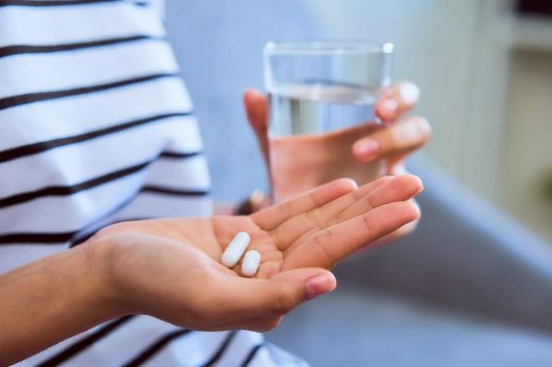 close up of a person's hand holding two small white capsules in the background is their other hand holding a glass of water