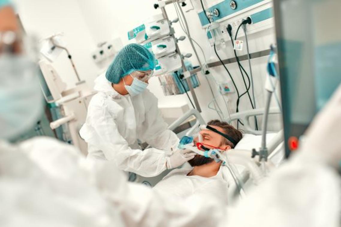 A person in a medical gown, mask and hair covering bends over a man in a bed surrounded by medical equipment.