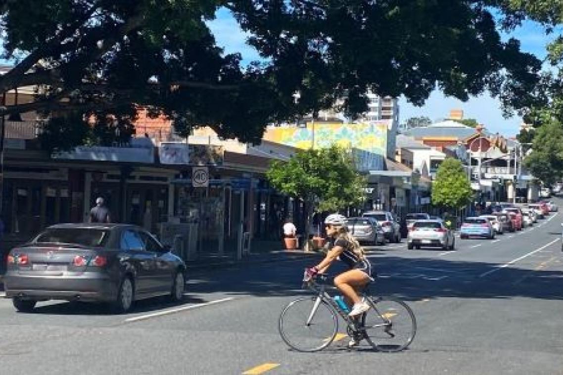 A cyclist rides across a busy road in the Brisbane suburb of West End, with a car passing in the other lane.