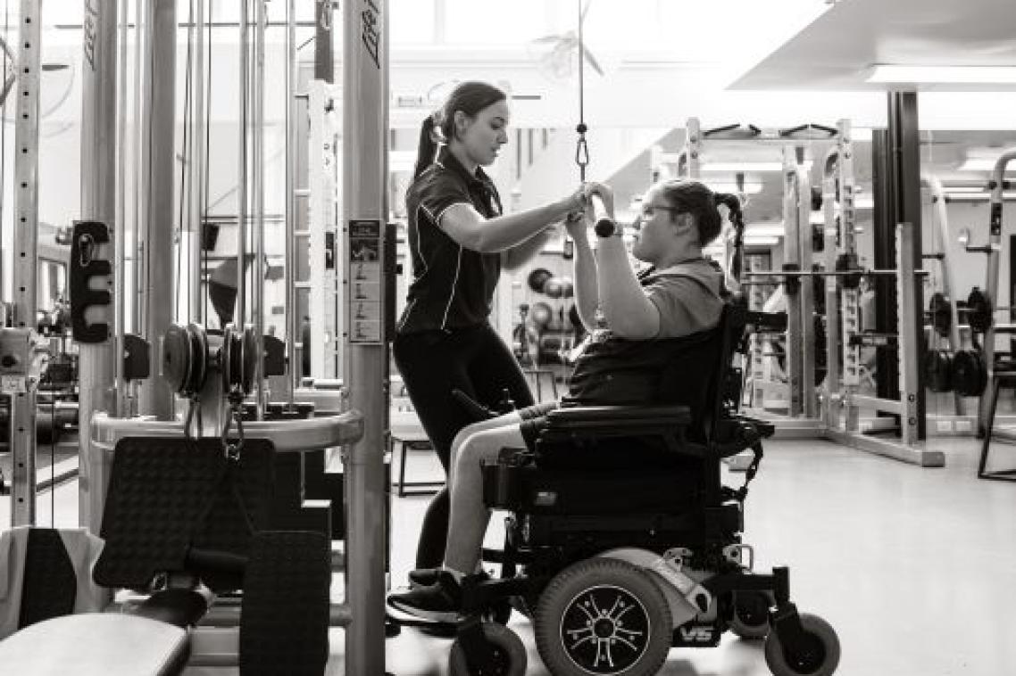 A young women seated in a wheelchair is working at a pull-down weights machine in a gym, assisted by a female coach. The photo is in black and white.
