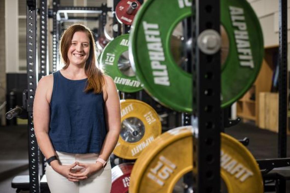 A woman stands smiling in a gymnasium, beside large colourful weights.
