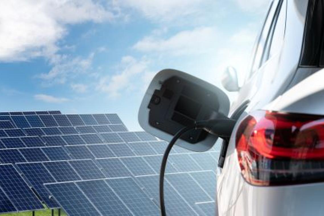 The side of an electric car, plugged in to a charging outlet with solar panels in the background.