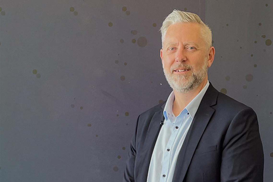 a man with a beard wearing a suit jacket stands in front of a grey wall