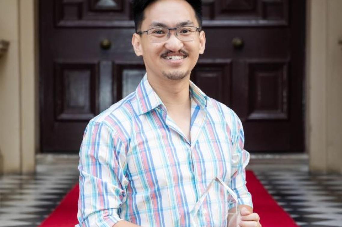 A man with black spiky hair and glasses smiles broadly outside double doors with a red carpet behind him. He is holding a glass trophy.