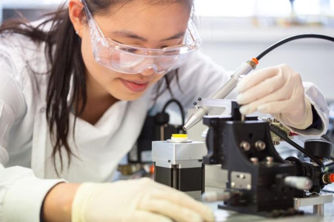 a woman in safety glasses looks closely at a yellow disk on a stand