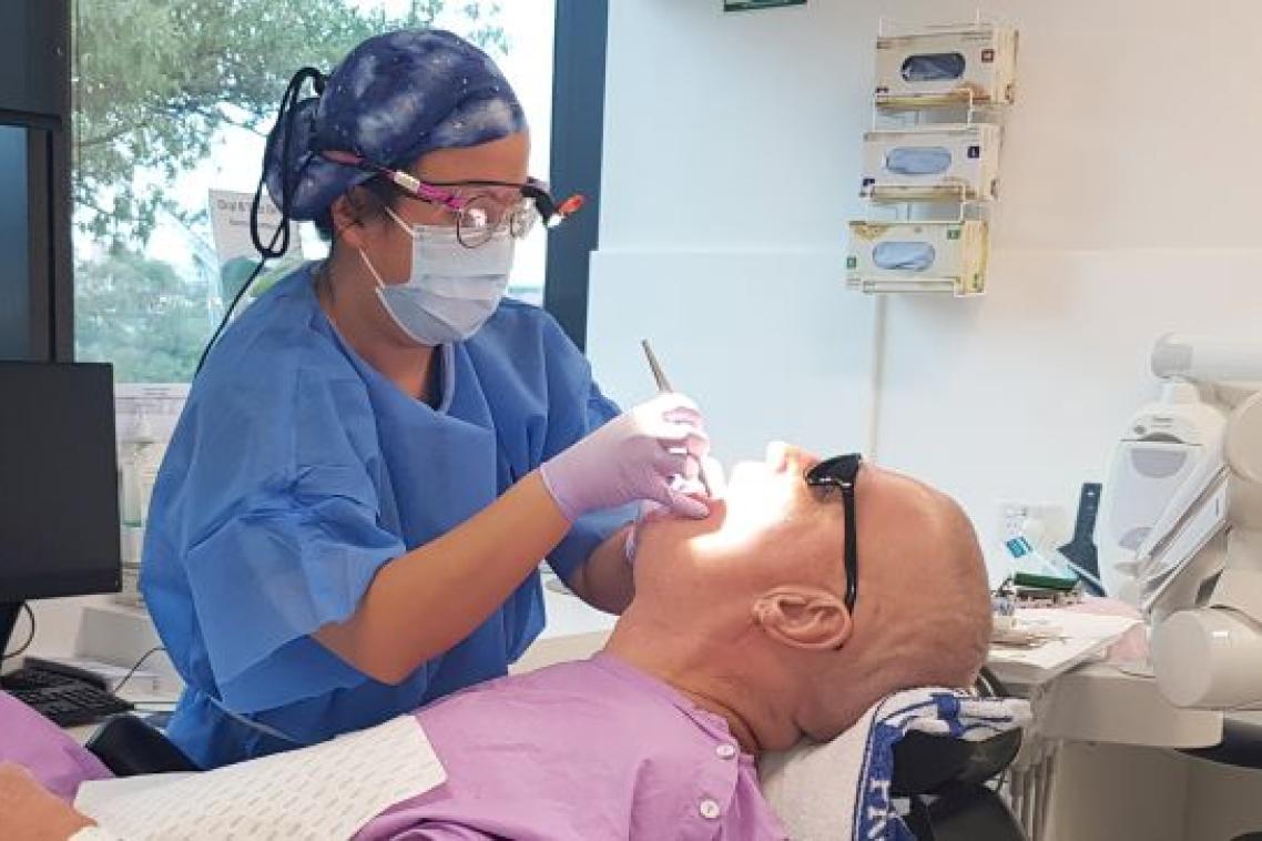 A person in a mask, safety glasses, gloves and hair net shines a light into the mouth of a prone man in pink clothing.