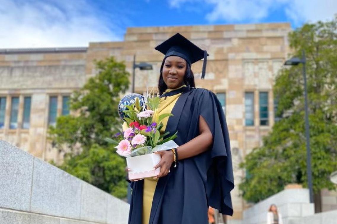 A young African woman stands in graduation cap and gown and holding flowers outside UQ's sandstone Great Court.