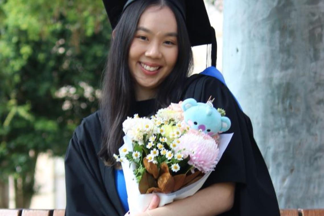 A young woman in graduation cap and gown sits on a bench seat in UQ's Great Court, clutching a bouquet of flowers with a small teddy bear inside it.