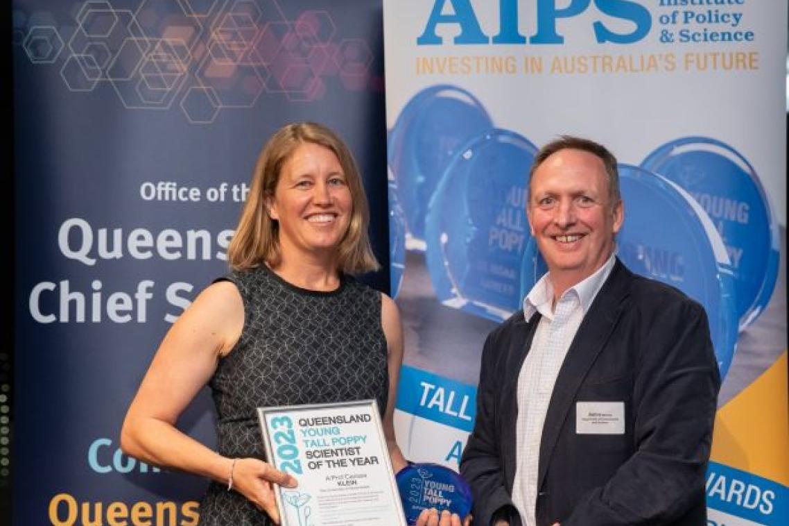Two people stand smiling in front of banners at an awards night, one of them is holding an award.
