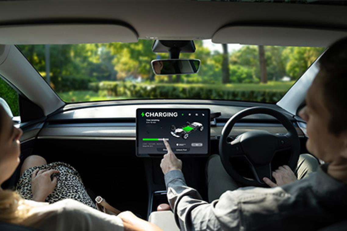 A photo taken inside an EV showing a driver and passenger in the front seat, the driver is pointing to the vehicle's charging screen.