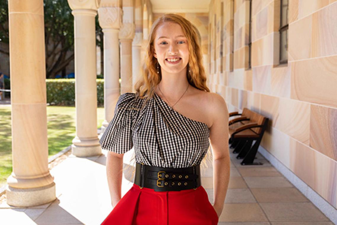 A young woman with long blonde hair standing under a sandstone walkway surrounded by columns