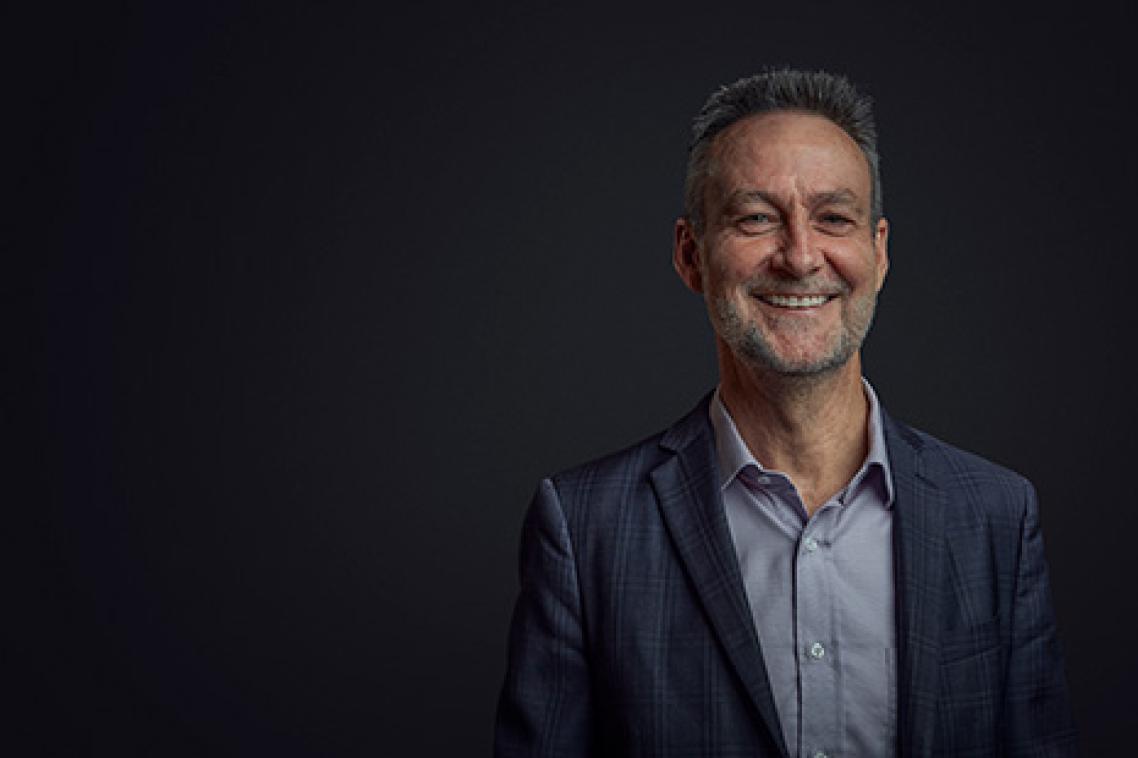 Glenn King wearing a jacket and collared shirt smiling broadly in front of a black background