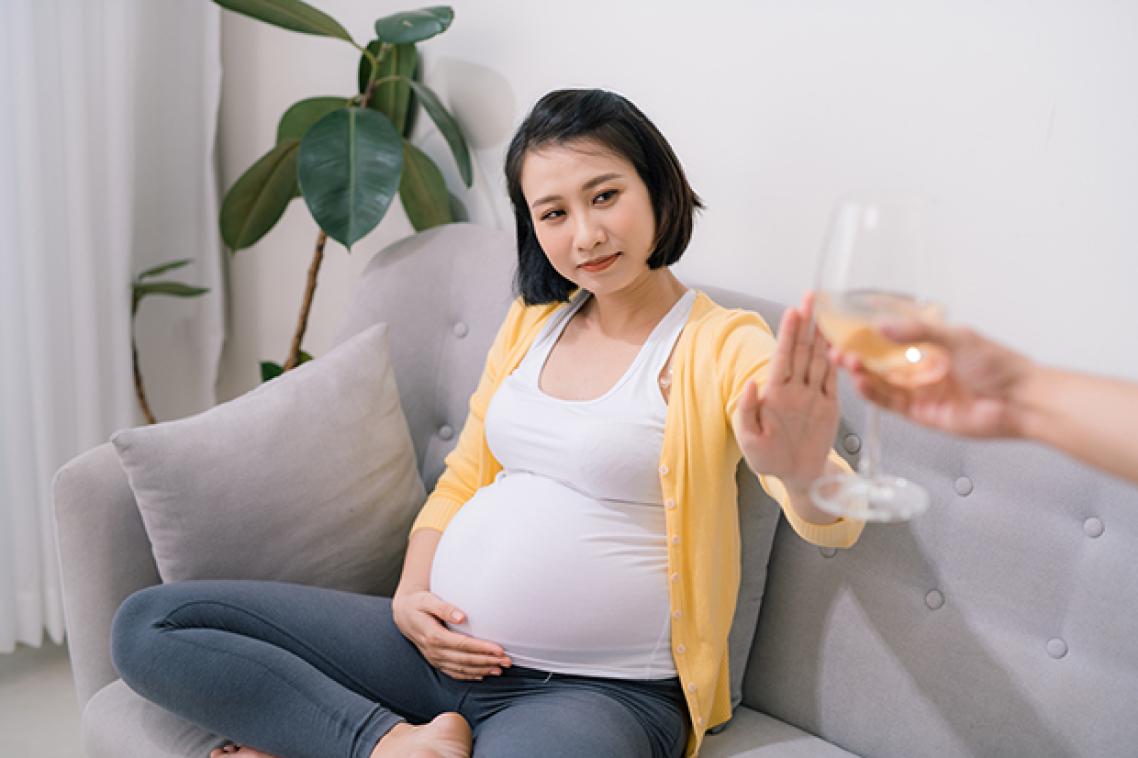 A pregnant woman sitting on a couch refusing an offered glass of wine