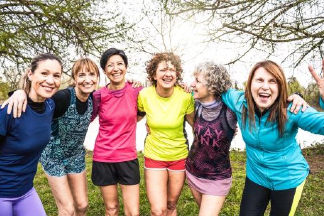 A line-up of six women standing outdoors dressed in exercise gear, smiling and with their arms around the person beside them.