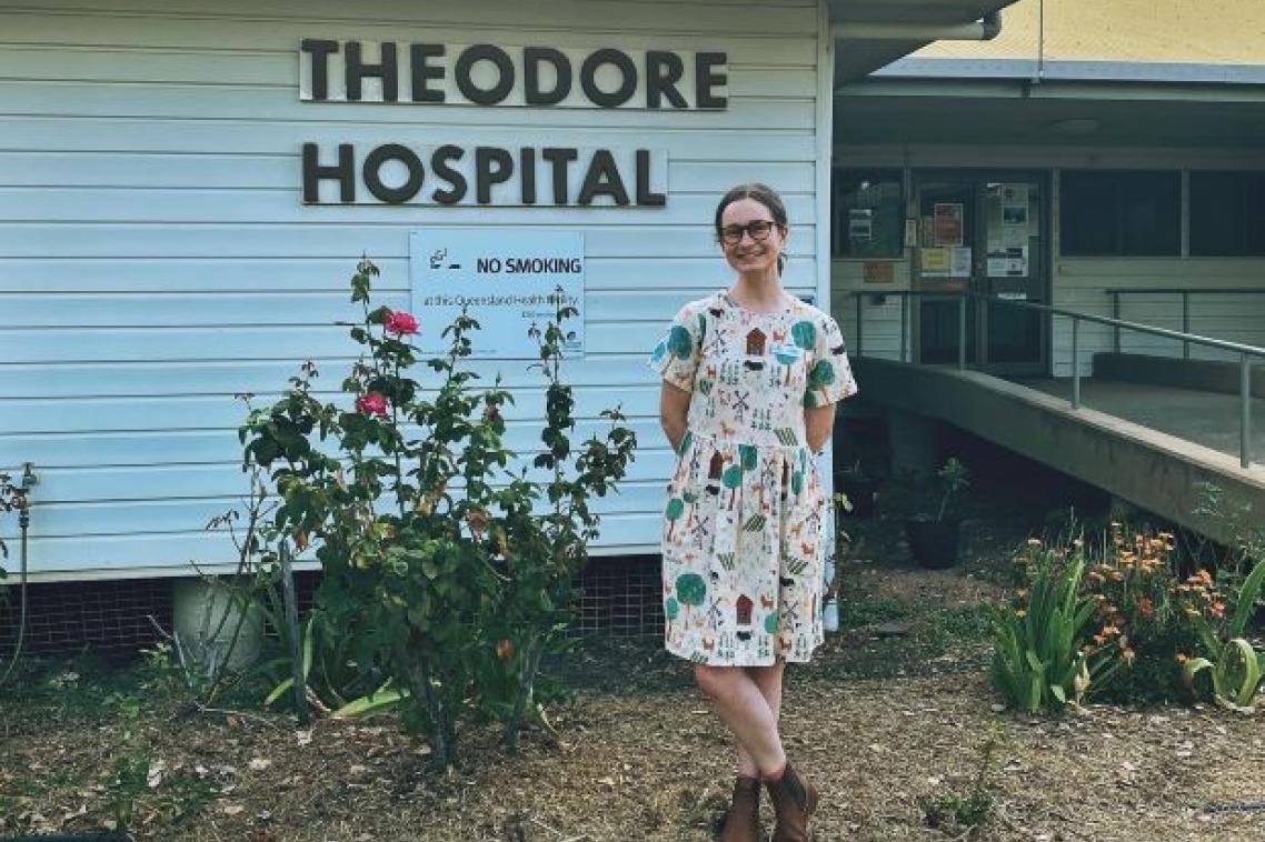 A woman in a dress and boots stands in front of a building which is labelled Theodore Hospital