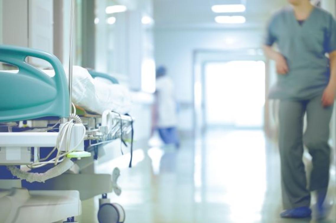 A hospital corridor with a trolley bed on one side, and an out of focus outline of a hospital worker in green scrubs on the other.