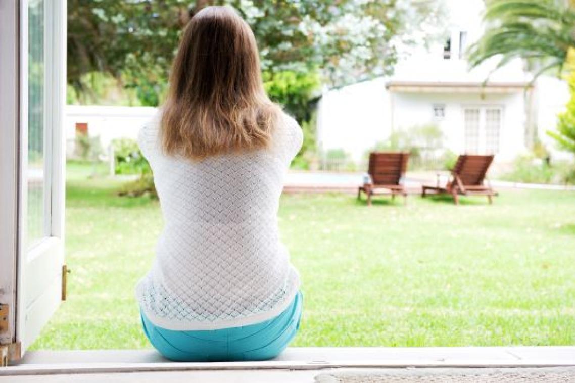 A woman sits at an open doorway with her back to the camera. She is facing a garden with a green lawn, trees and wooden chairs.
