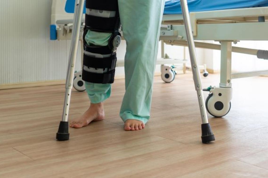 The legs of a person by a hospital bed in green clothing with a braced knee and crutches