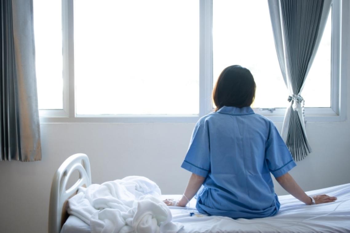 woman in blue shirt sits on a bed and looks out of a window
