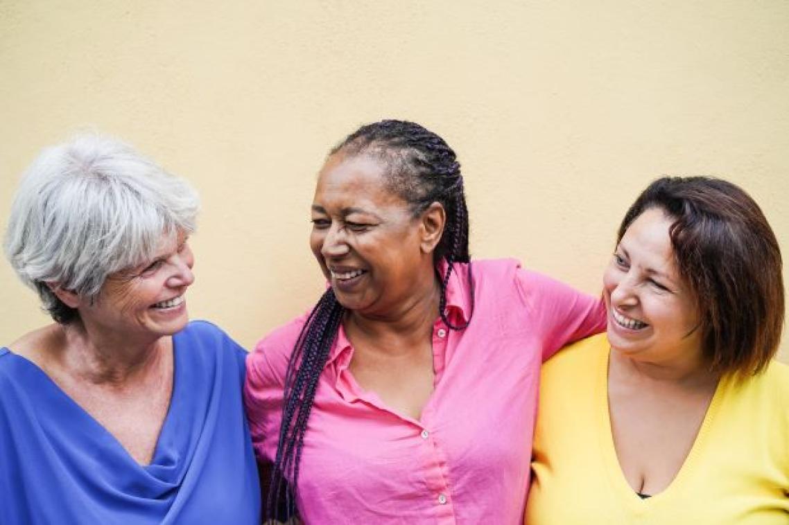 Three women in brightly coloured shirts smile and laugh. They have their arms around each other.