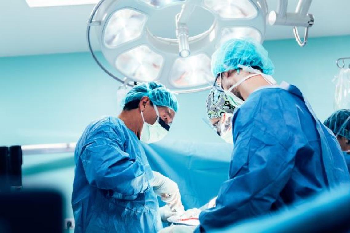three people in blue gowns, surgical masks and gloves stand under a bright light looking down