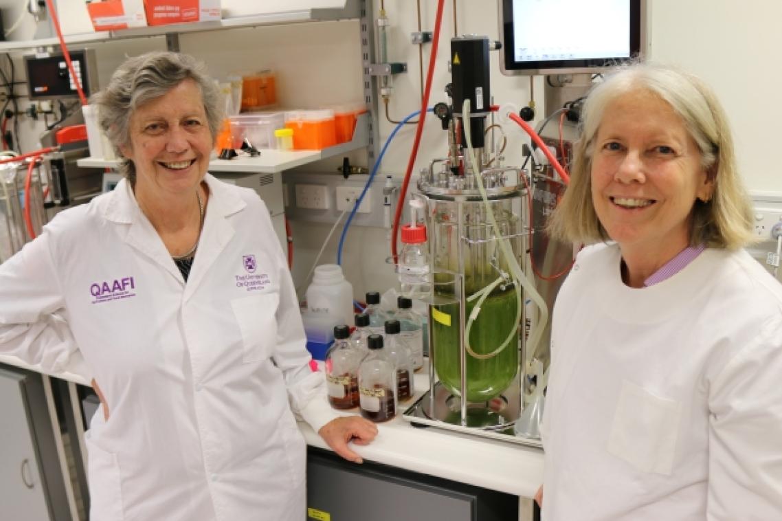 two women in white coats smile in a laboratory in front of a device containing green liquid
