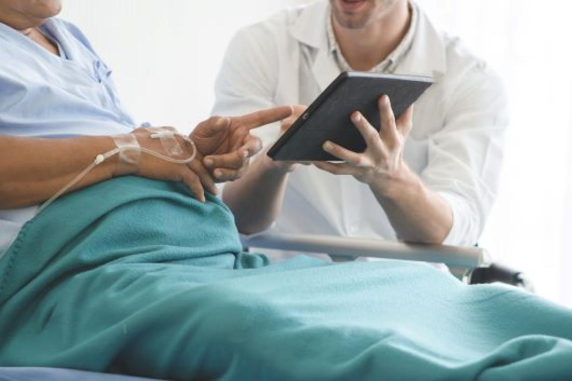 A patient sits up in a hospital bed and points at a tablet being held by a clinician sitting beside them.