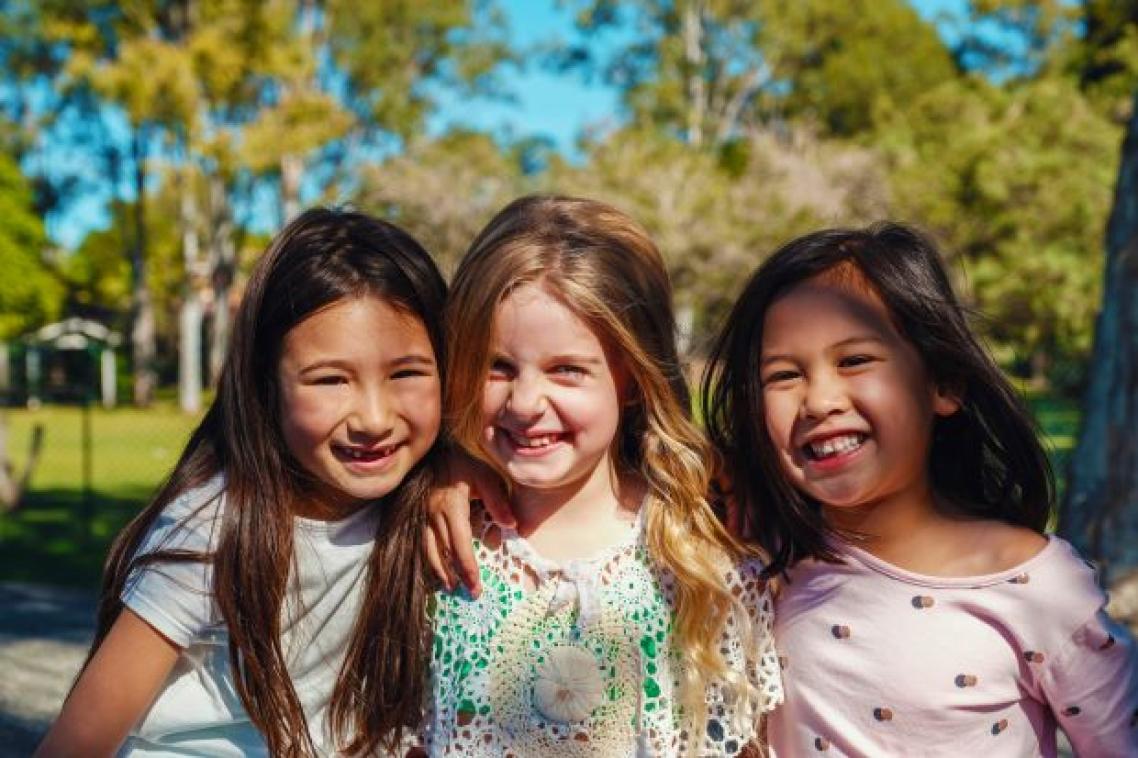 three smiling girls with long hair in the sunshine there is grass and trees behind them