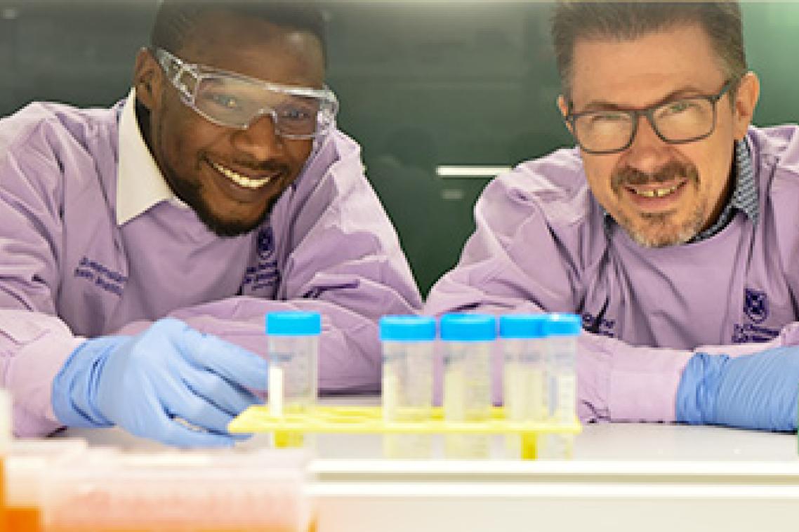 two men in purple lab coats and wearing glasses look at a rack of blue-lidded sample pots