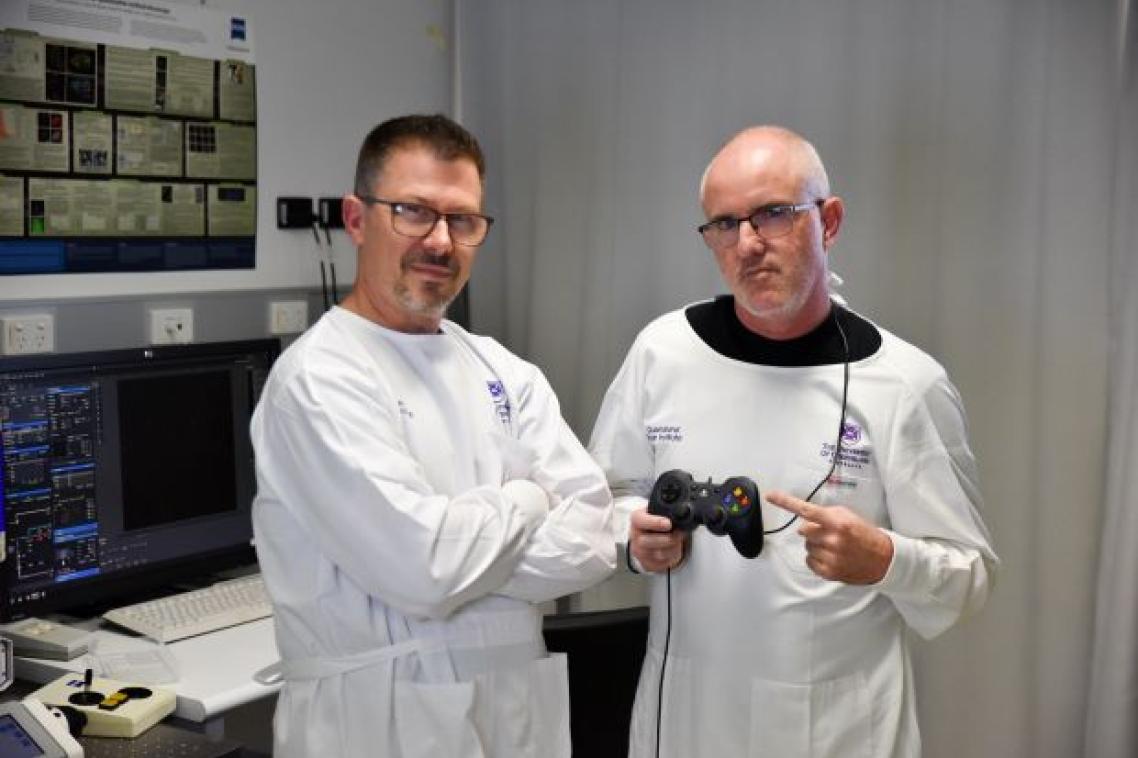 Two men in white lab coats in an office look at the camera, one holds a gaming controller.