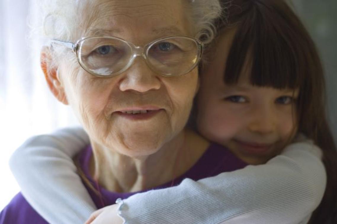 An older woman with white hair and glasses looks at the viewer. A girl of around 5-6 years old has her arms around her neck affectionately.