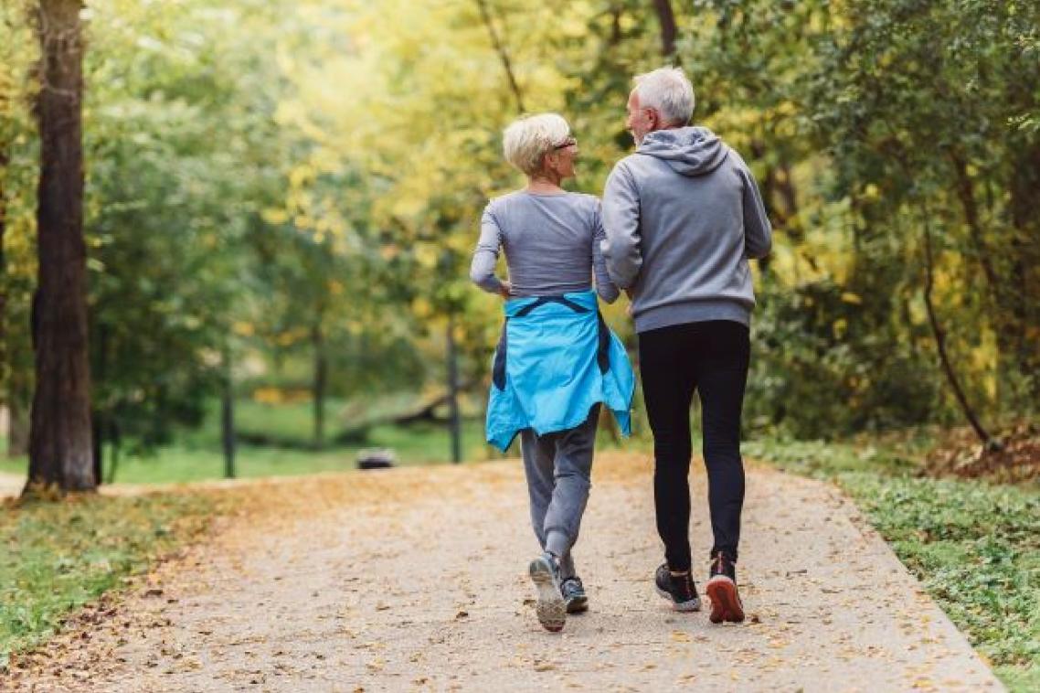 two people with grey hair are walking away from the camera along a sandy track with green vegetation either side