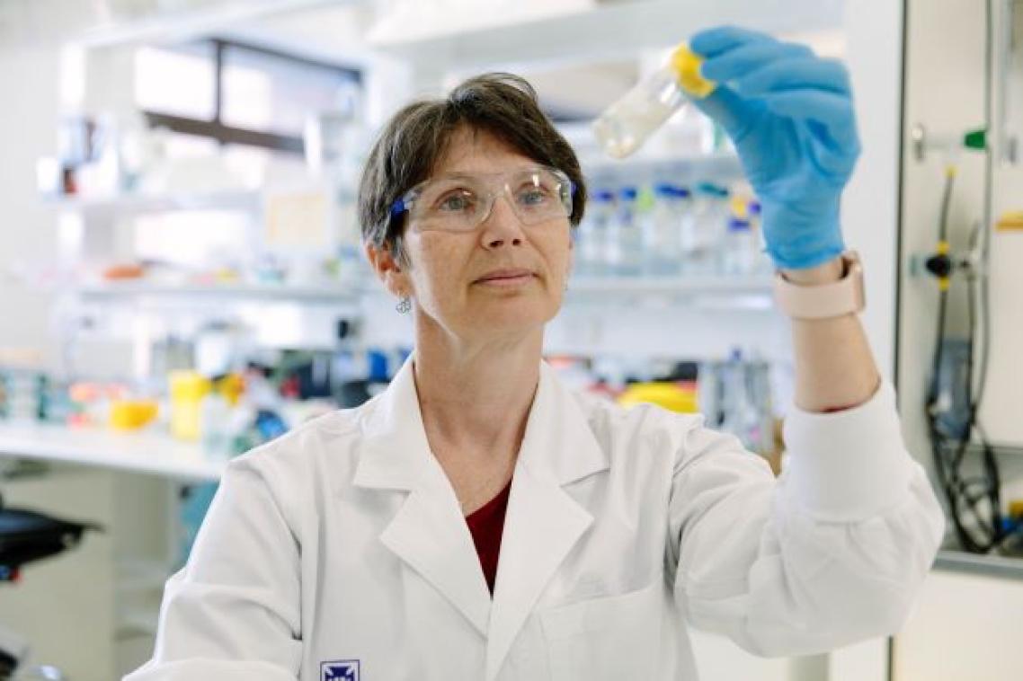 A woman stands in a laboratory wearing a white coat and glasses and holds up a glass vial