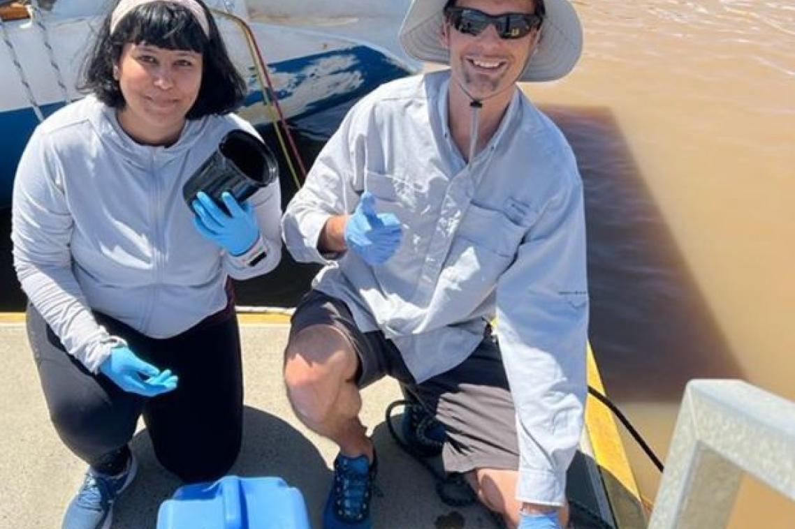 Two people on a dock collecting water samples with a boat and brown river behind them.