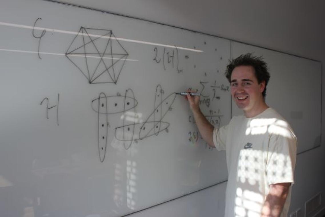 A man stands at a whiteboard, writing a mathematical equation. He is smiling at the camera.