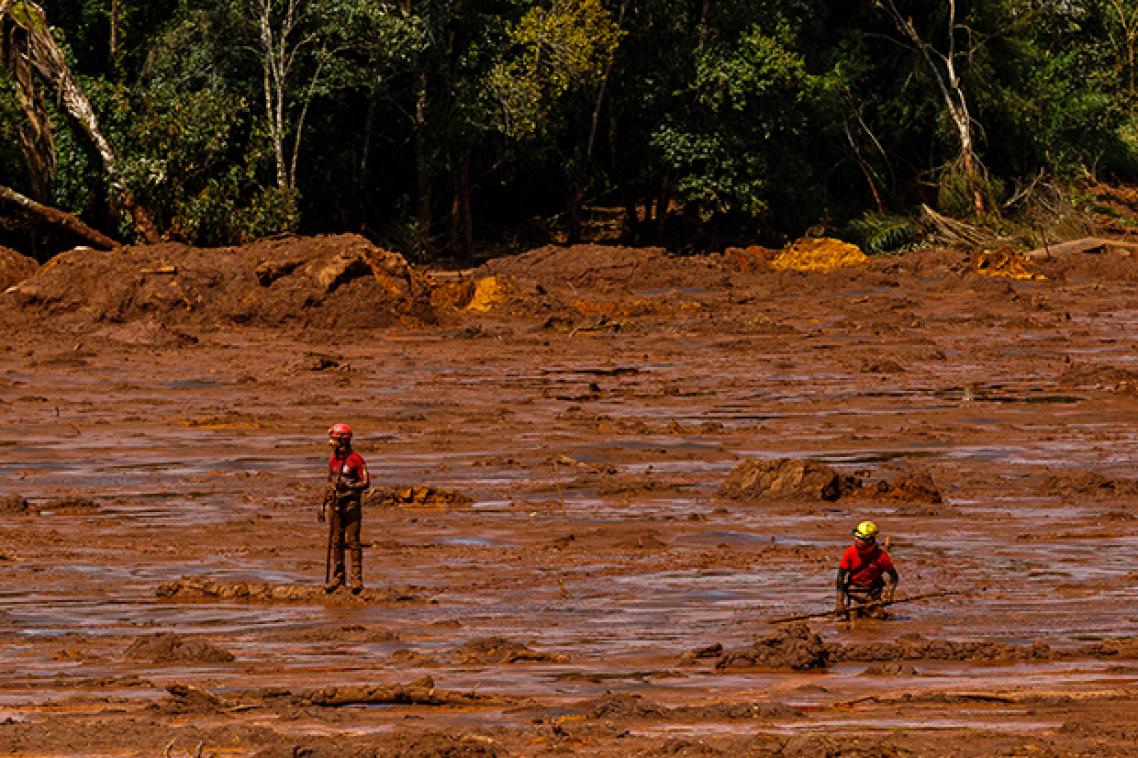 Two people in red shirts and helmets stand in red mud, there are trees in the background
