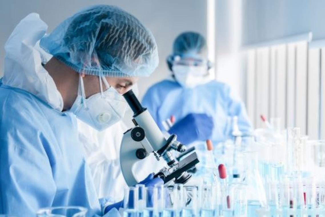 A scientist in cap, gown and mask stands in profile looking down through a microscope as a second scientist looks on in the background. They are in a lab.