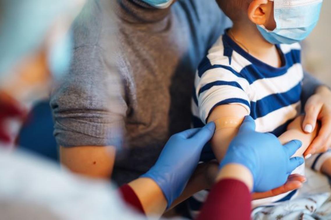 Gloved hands are smoothing a bandaid onto a child's upper arm. The child is on the lap of an adult carer.