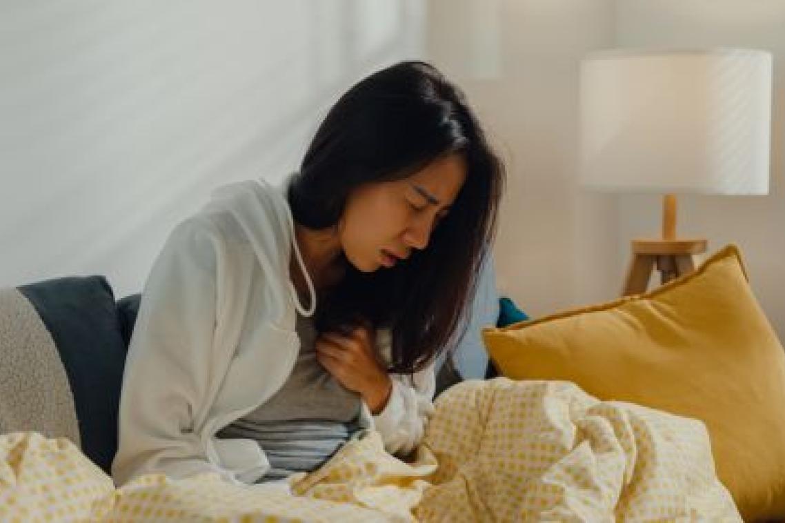 A woman with long black hair is sitting up in bed wincing in pain and one hand is on her chest.