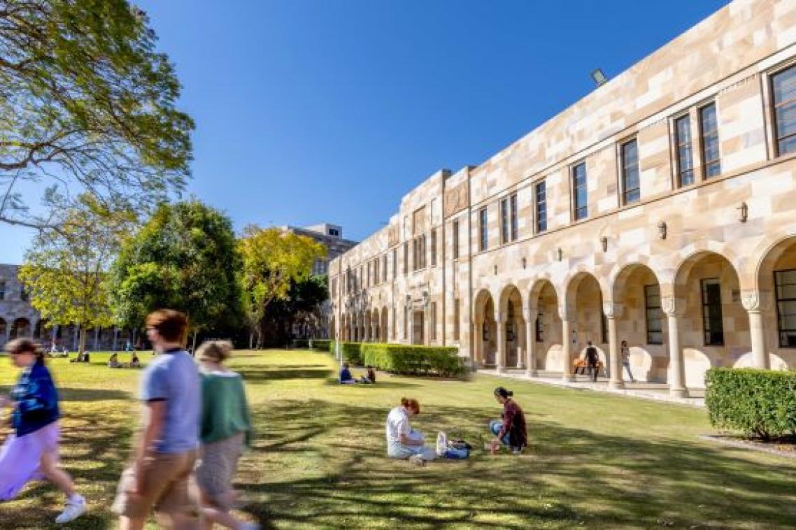 people walk and sit on grass by stone buildings with an arched walkway