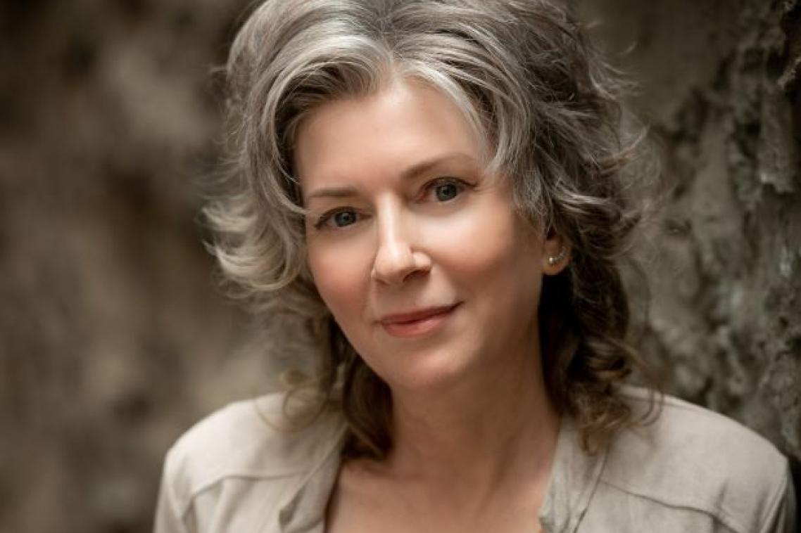 A woman with grey hair wearing a cream jacket over a rust coloured shirt.