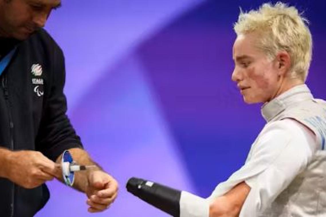 A female para athlete watches as her coach approaches with a prosthesis for her fencing competition. 