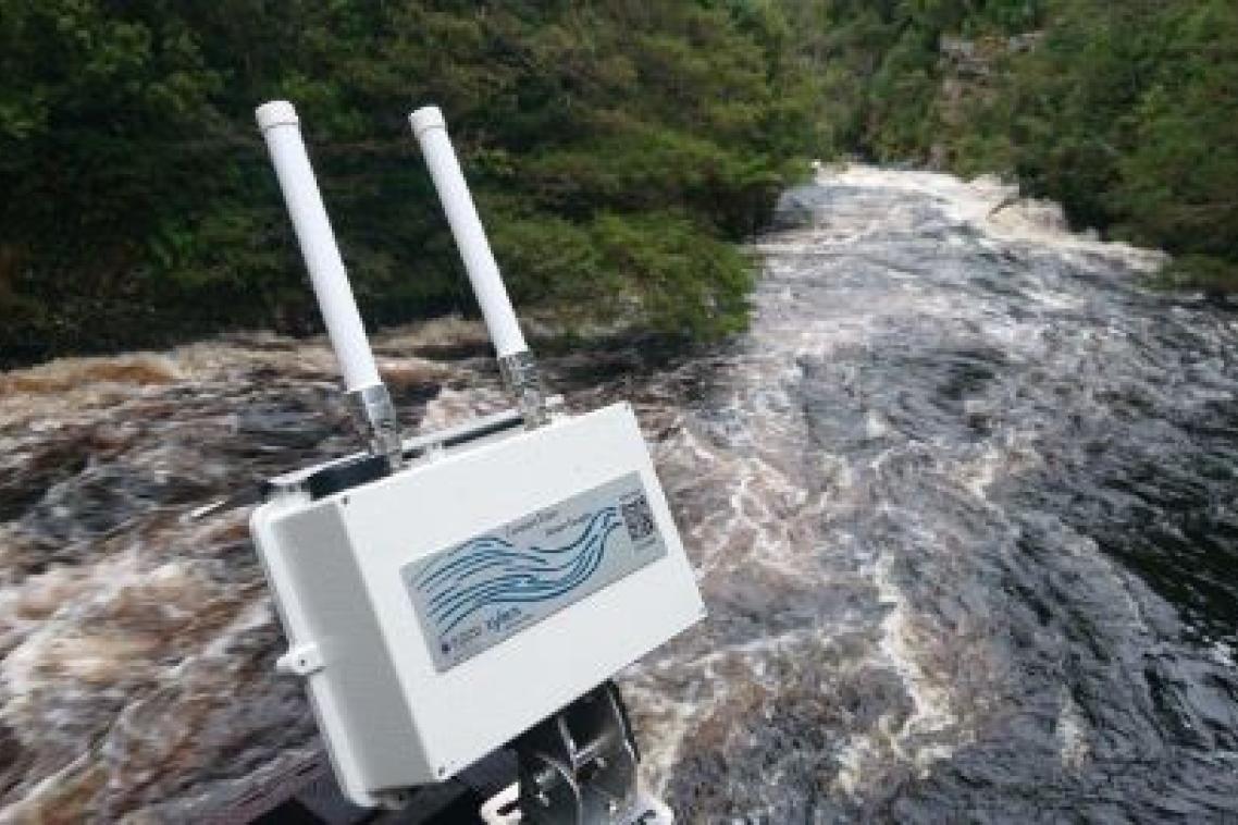 A white camera-looking object with two antennas sits above fast flowing brown water with bushland in the background.