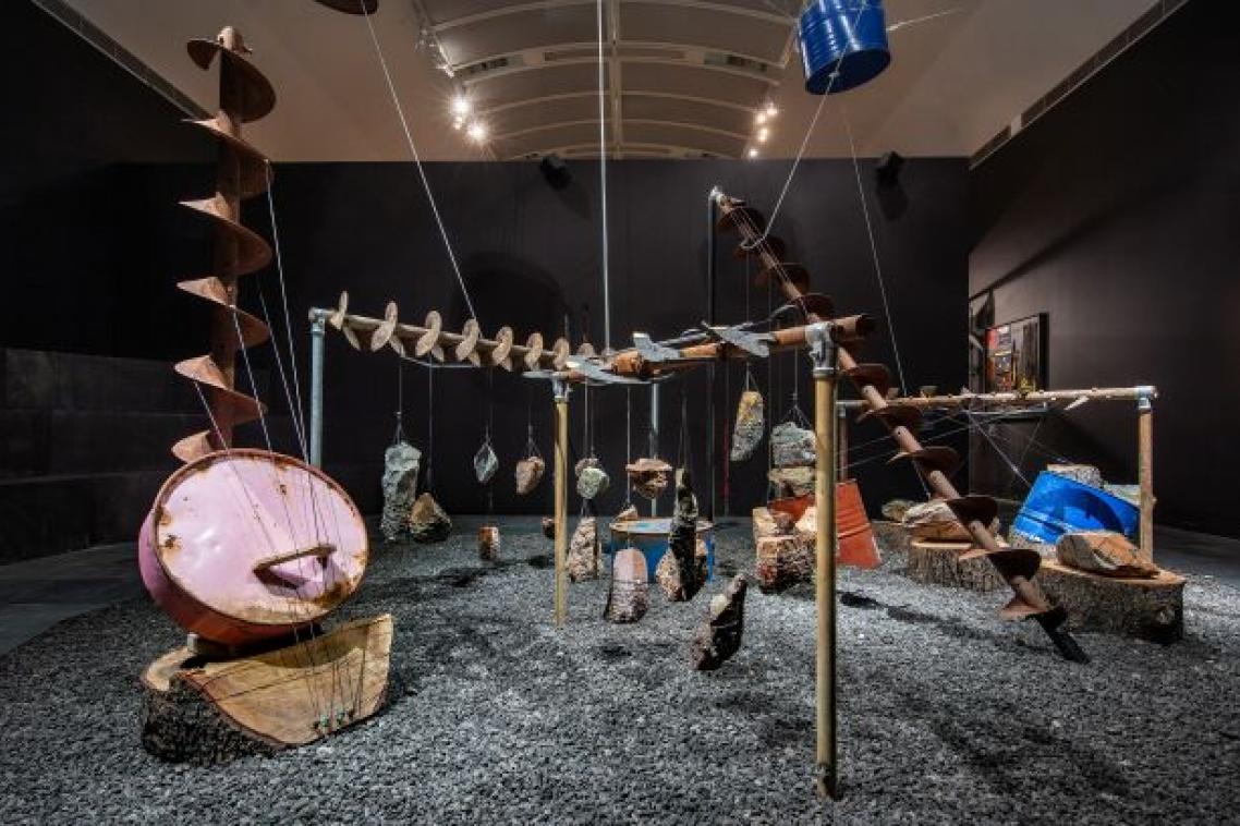 A room with dark walls is filled with an art installation of industrial mining equipment repurposed into musical instruments.
